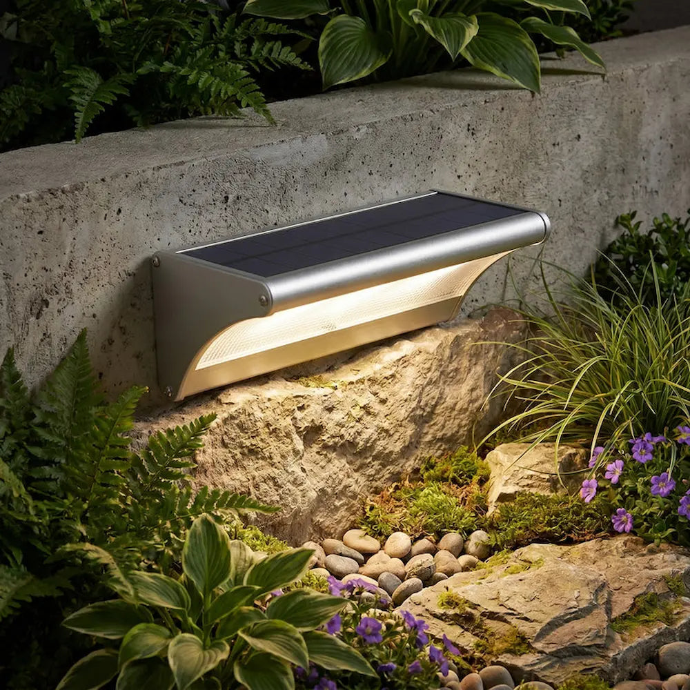 Solar-powered outdoor light fixture on a stone wall with plants and flowers in the background