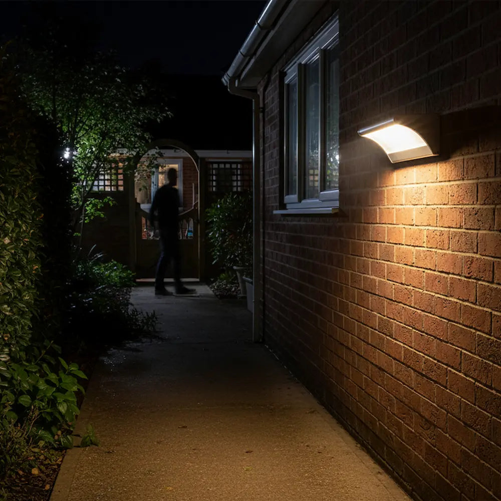 Person walking down a dark pathway illuminated by a wall-mounted light fixture on a brick wall.