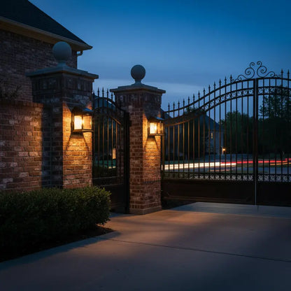 Gated entrance to a residential area at night with illuminated brick pillars and wrought iron gate.